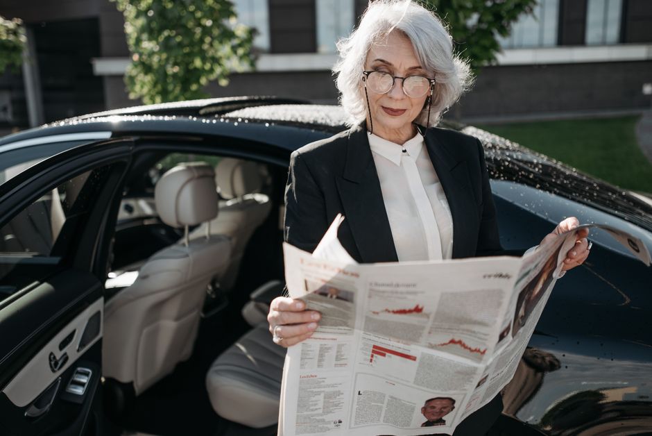Jornalismo Digital — Elegant senior woman in formal attire reading a newspaper next to a luxury car outdoors.